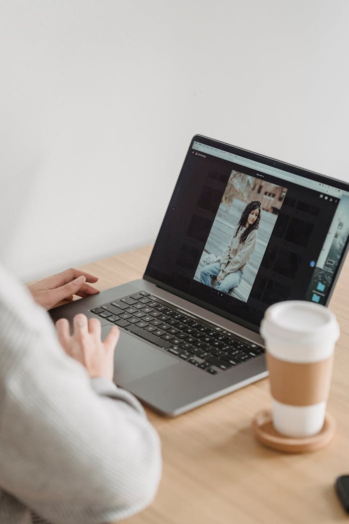 Crop unrecognizable person looking at image on netbook screen while sitting at desk with takeaway coffee